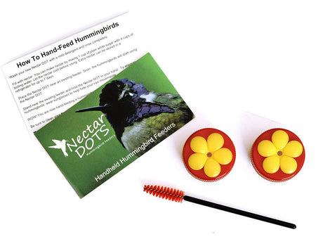 Product photo showing two red and yellow Nectar DOT hummingbird feeders with a cleaning brush and instruction leaflet featuring a vibrant hummingbird illustration