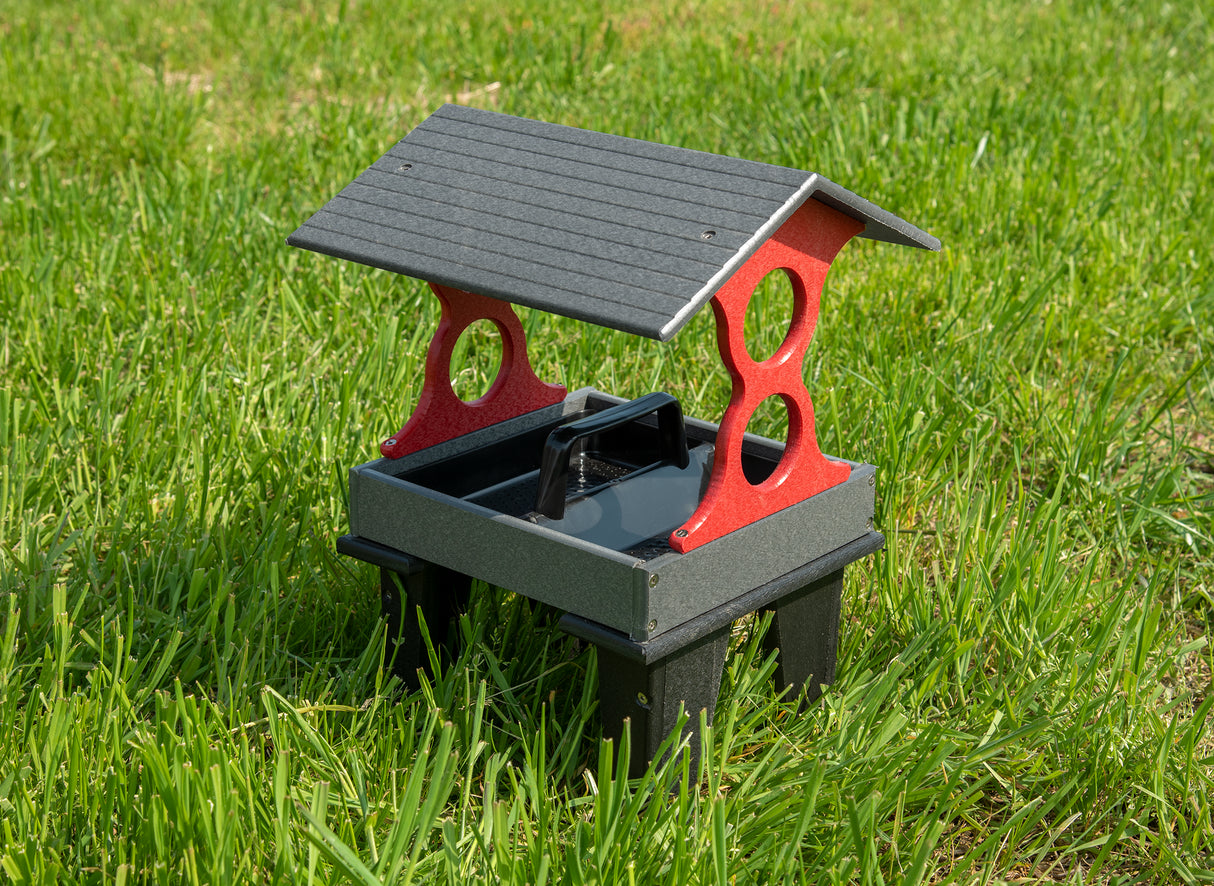 Plastic bird feeder in red and gray on green grass featuring a sloped roof and a central carrying handle with side cutouts