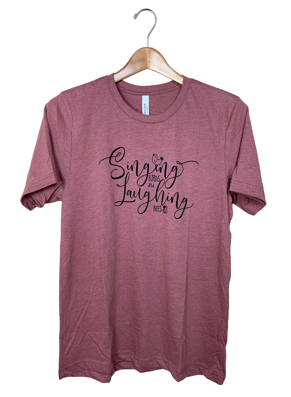 Canvas Bella+Canvas t-shirt featuring a dusty rose color a stylish cursive design that says Singing Birds and Bees with small bird and bee icons in black ink on a wooden hanger