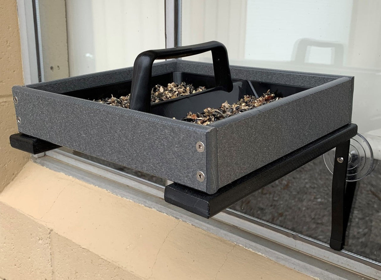 Photograph of a gray plastic window bird feeder with black accents and a handle containing mixed birdseed attached to a window with suction cups showing visible screws and a textured surface