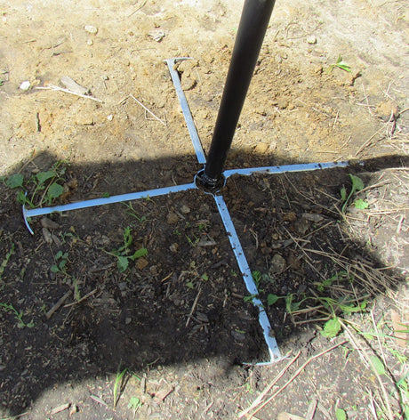 Photograph showing a black metal pole base with four light gray metal stabilizing arms set in dark brown soil featuring small green plants and a distinct shadow