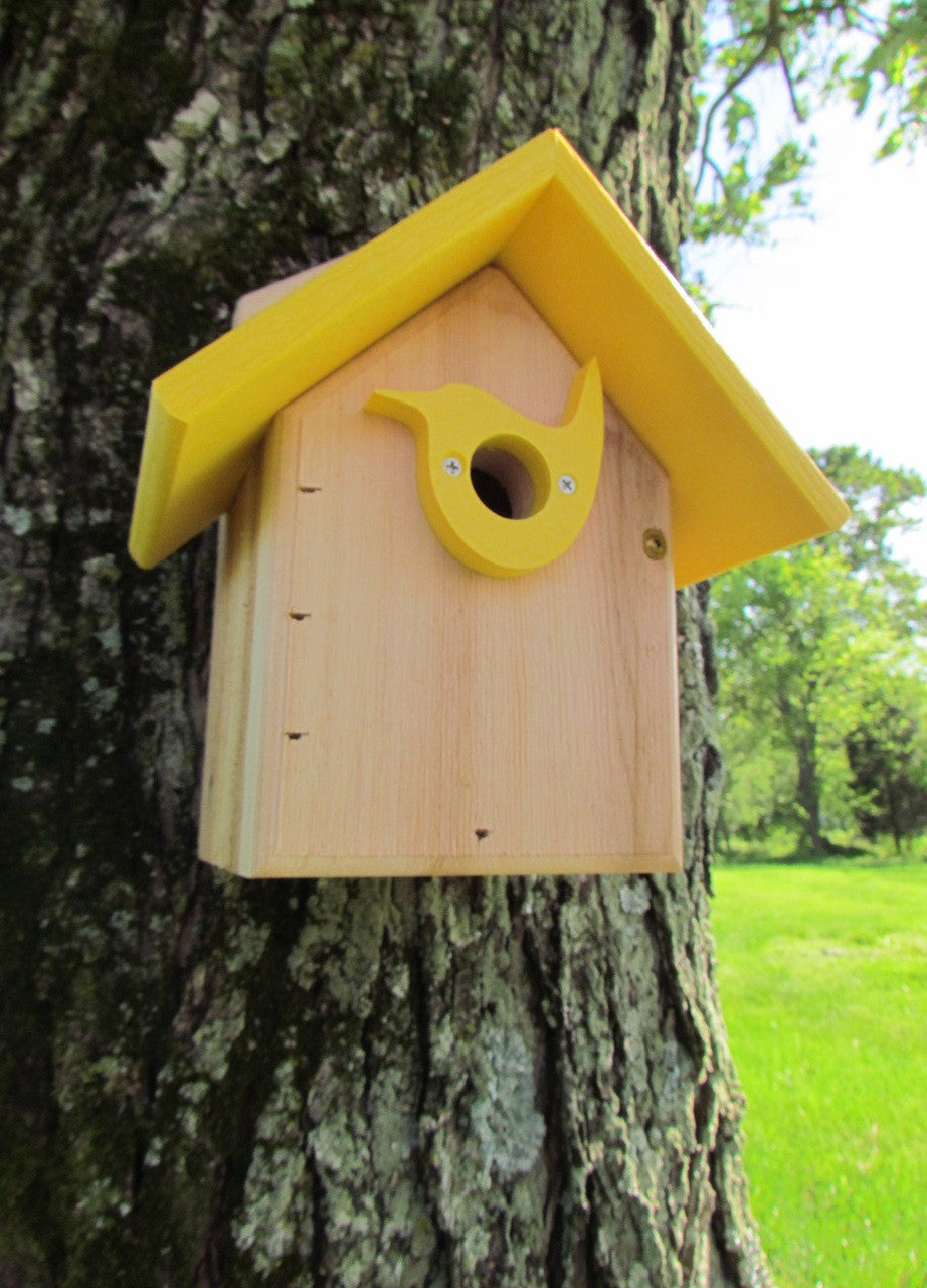 Wooden birdhouse in a backyard setting attached to a tree featuring a yellow bird-shaped entrance bright yellow roof and visible wood grain.