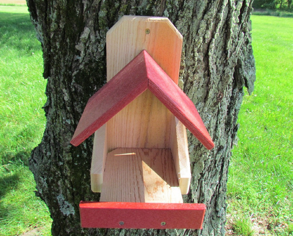 Handcrafted wooden bird feeder with red roof and perch against a tree in a grassy field showcasing vibrant red accents and natural wood grain