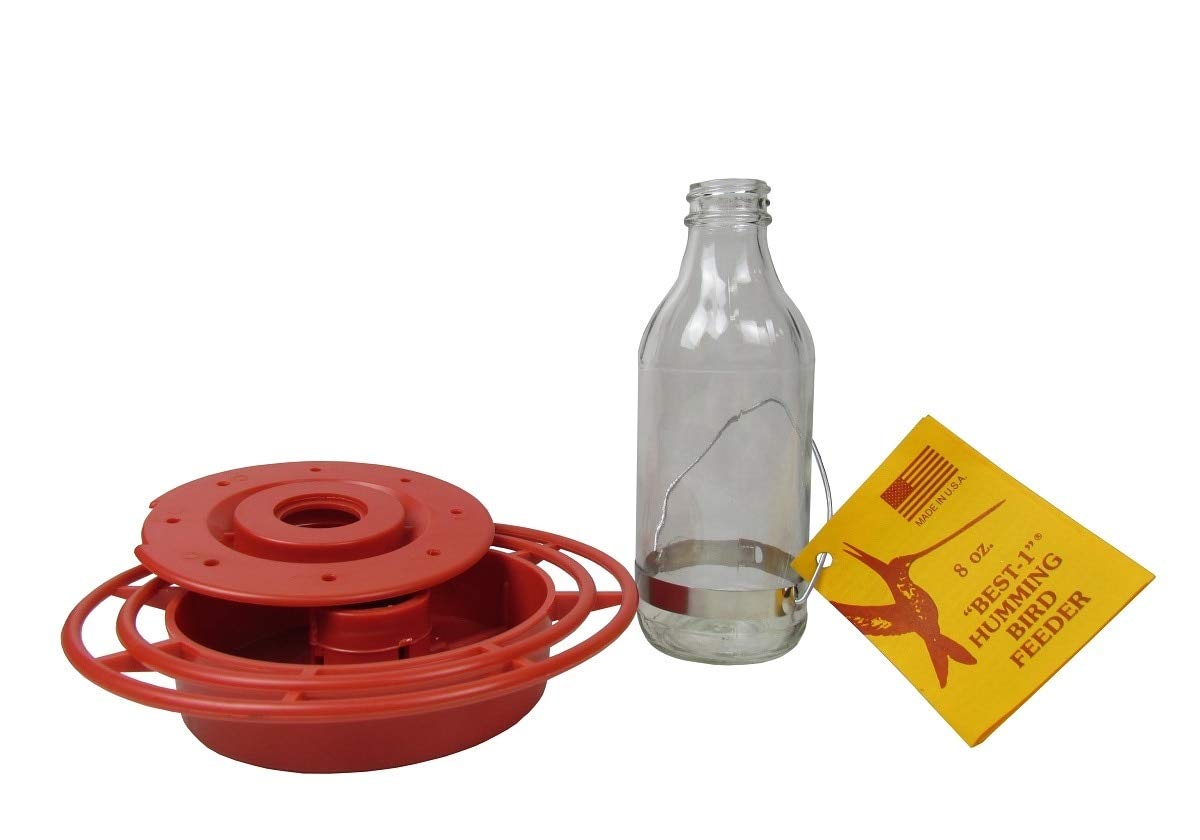 Product shot showing a Best-1 hummingbird feeder featuring a red plastic base, a clear glass bottle, and a yellow tag with hummingbird graphic and text details