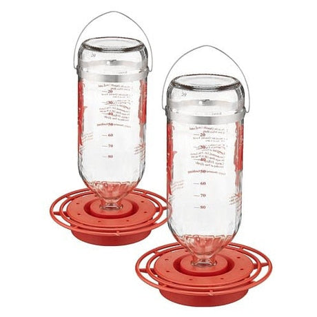 Two glass hummingbird feeders with red plastic bases and wire handles showing red measurement markings on the glass