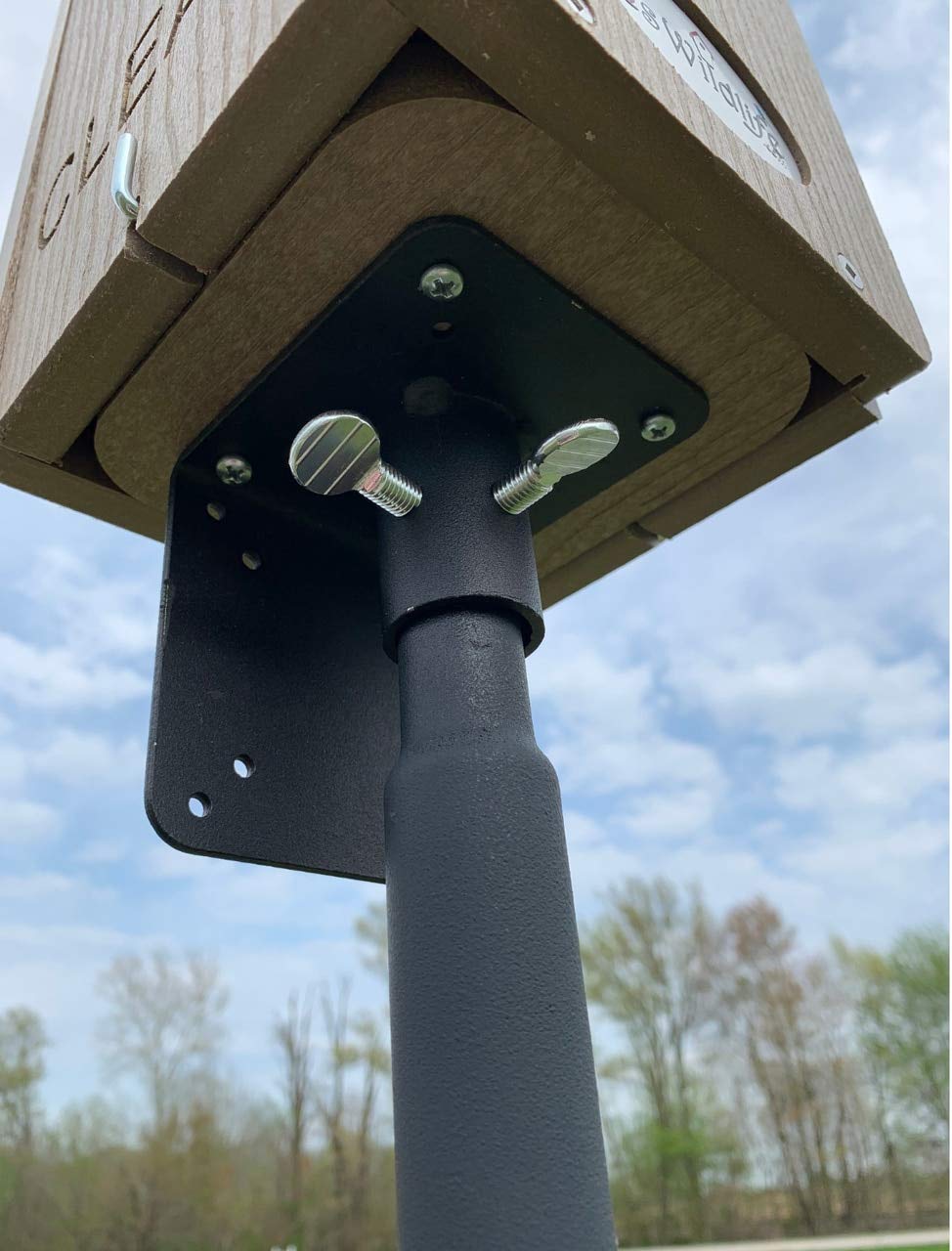Close-up photograph of a wooden birdhouse mounted on a black metal post featuring silver wing nuts and a textured surface against a backdrop of trees and a partly cloudy sky