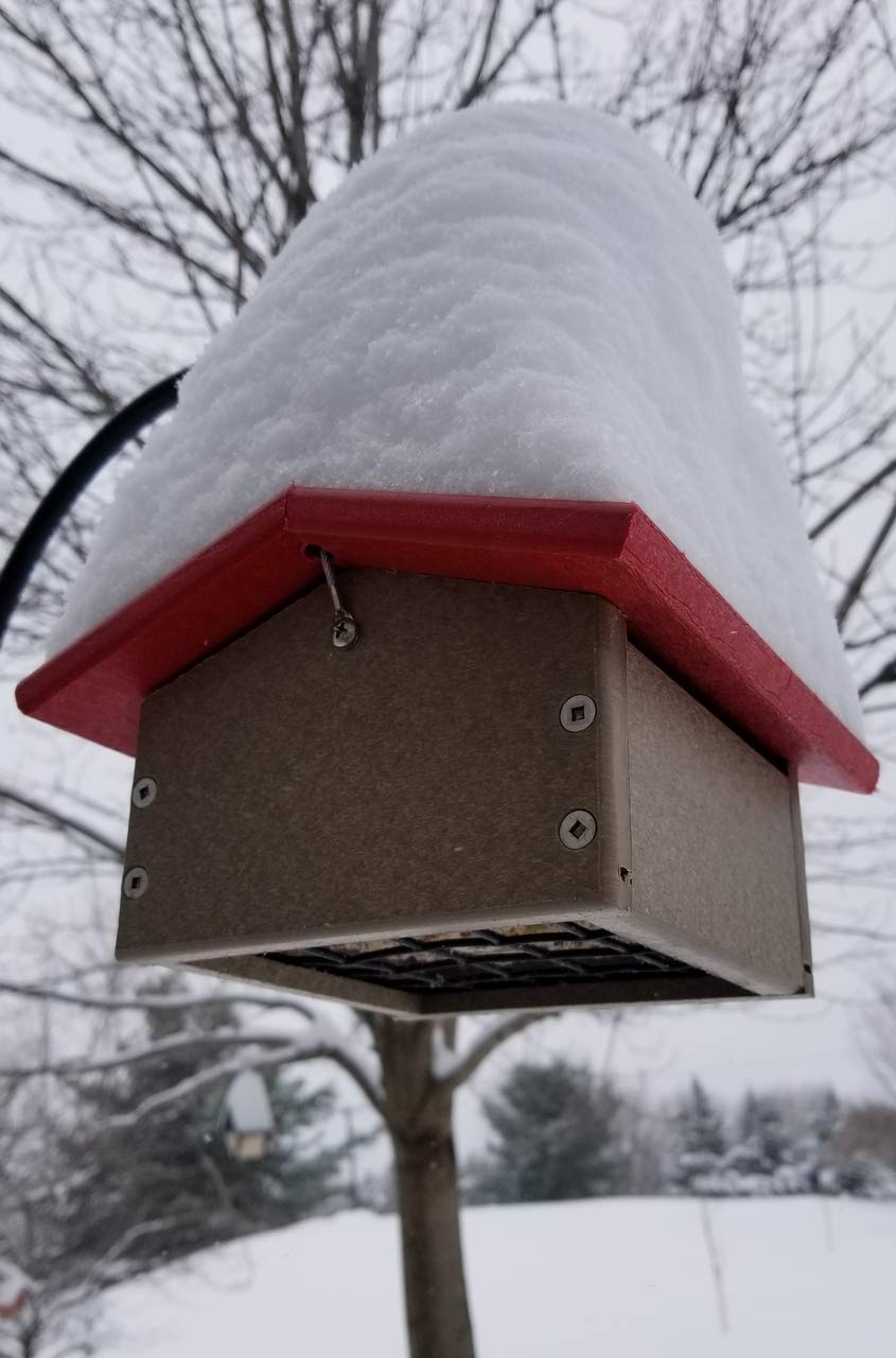 Photograph of a snow-covered bird feeder with a red roof and taupe body featuring visible screws and a wire hanger in a snowy backyard with a tree and another smaller bird feeder in the background