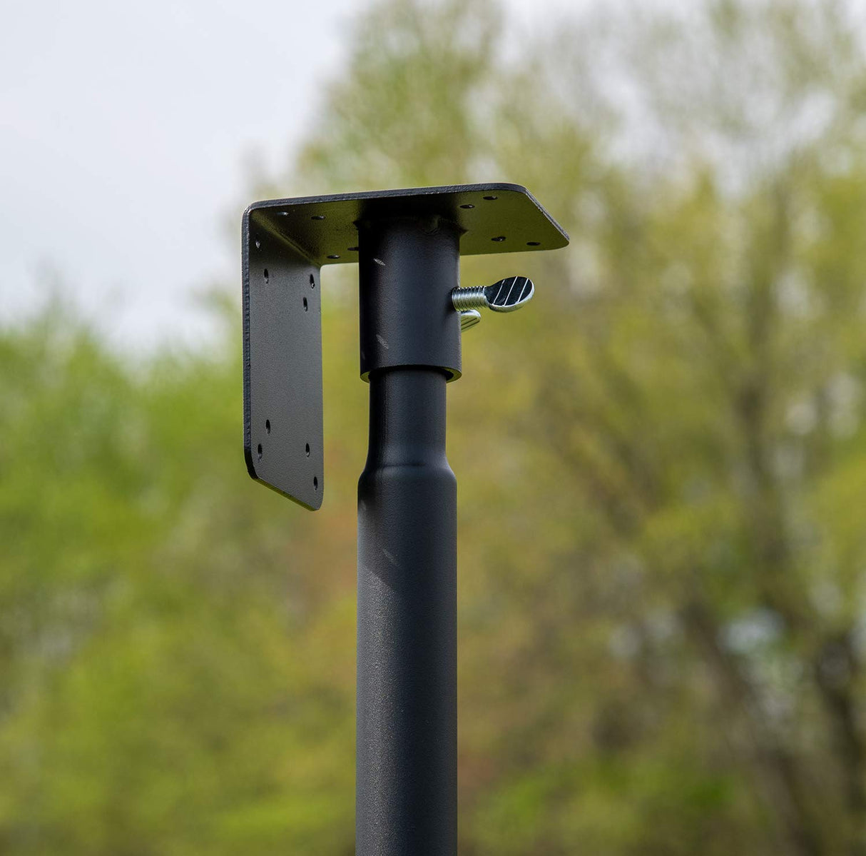 Close-up photo of a black metal post with a square mounting plate and a silver thumb screw against a blurred background of green trees