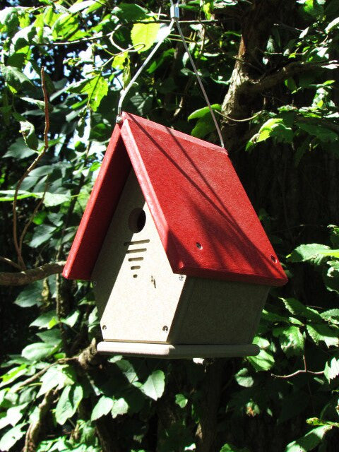 Recycled plastic birdhouse hanging in a lush green garden featuring a vibrant red roof and beige body showcasing ventilation slots and visible screws