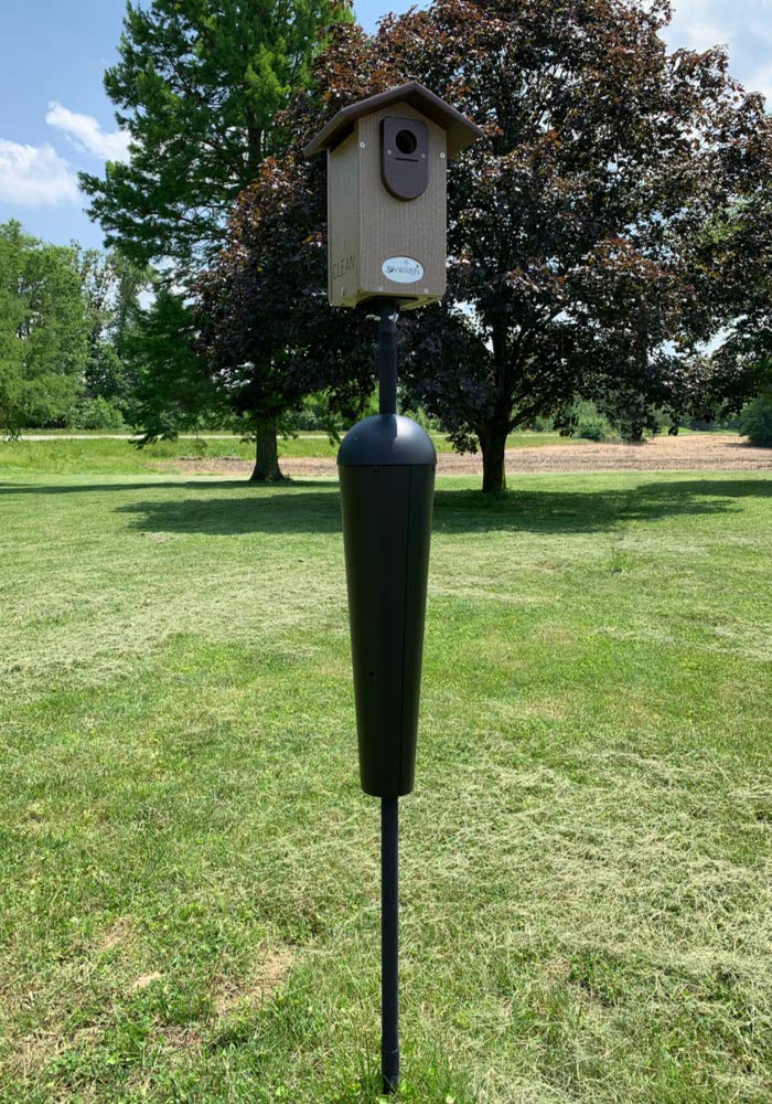Photograph of a Brown Birdhouse mounted on a black pole with a predator guard in a grassy field near green trees and a distant horizon
