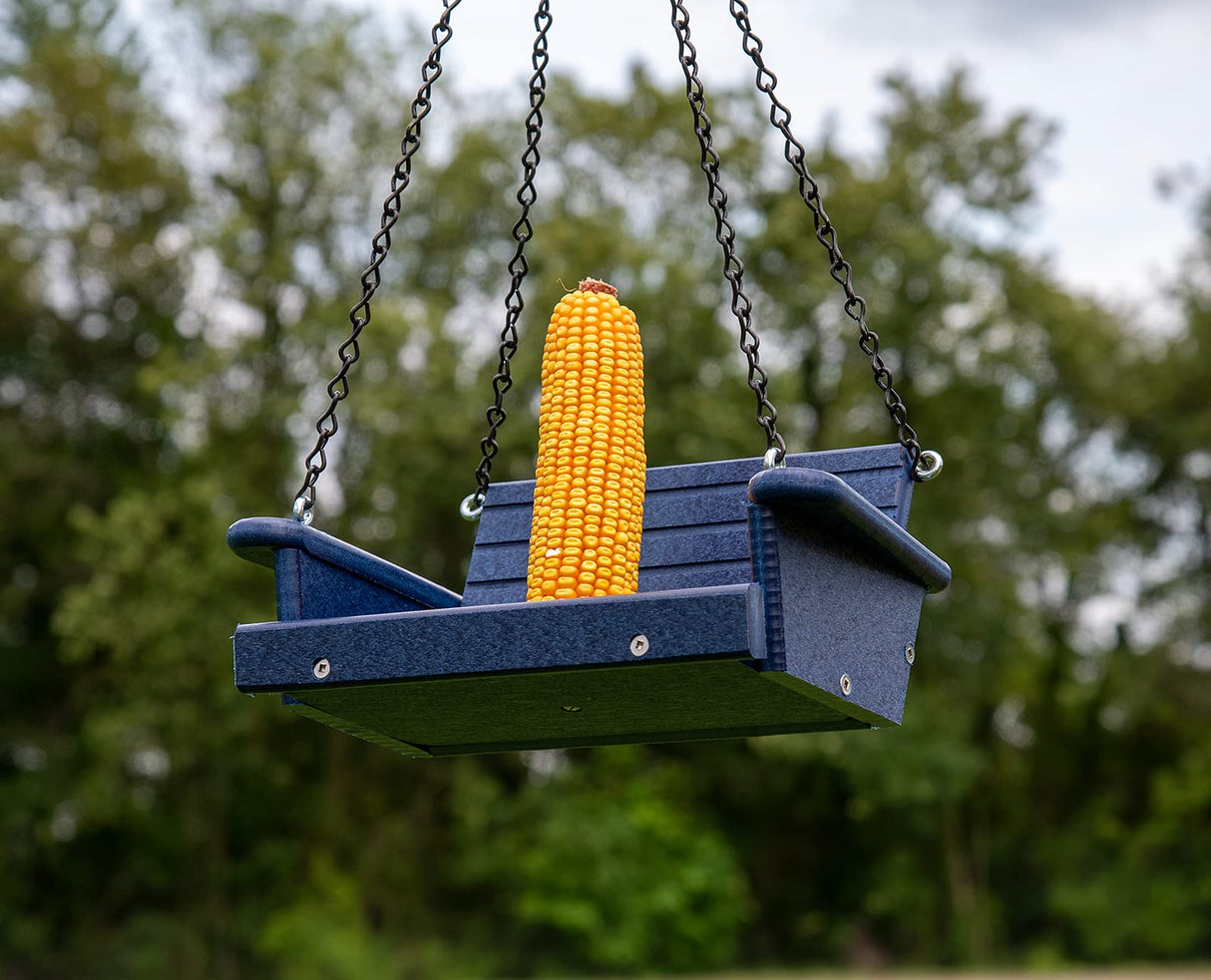 Photograph of a blue plastic bird feeder shaped like a swing outdoors with a bright yellow ear of corn and dark metal chains hanging amidst a lush green background of trees