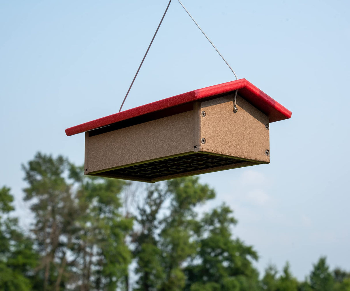 Recycled plastic bird feeder hanging outdoors against a backdrop of lush green trees showing a brown body bright red roof and visible screws