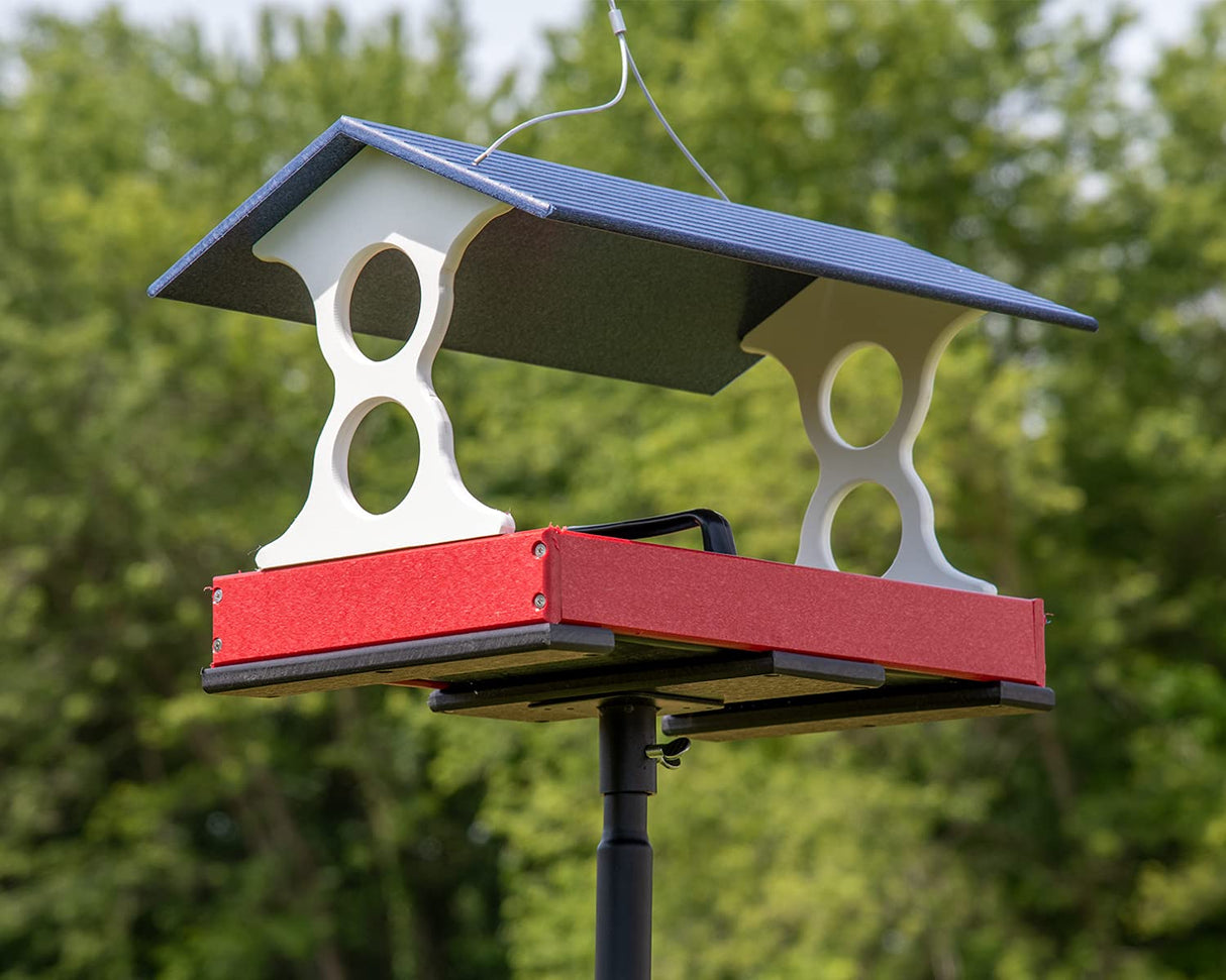Plastic bird feeder with red tray, white supports, and a blue roof outdoors against a green leafy background