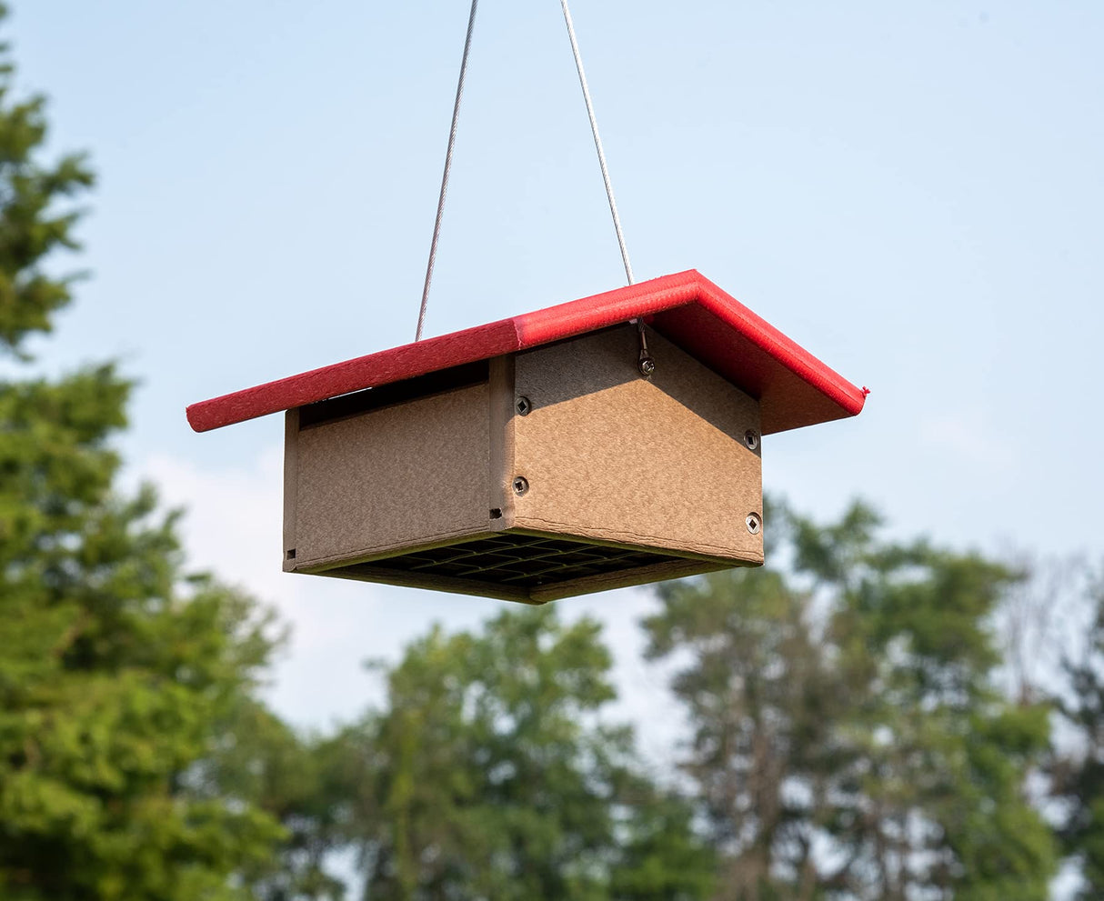 Recycled plastic bird feeder hanging outdoors against a backdrop of lush green trees featuring a bright red roof tan body and a wire mesh floor
