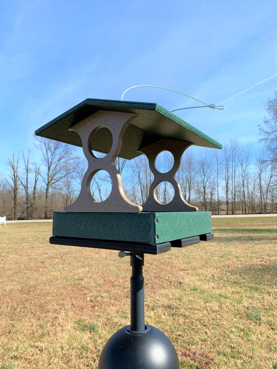 Recycled plastic bird feeder in a rural setting featuring a dark green roof taupe accents and a black weighted base against a backdrop of trees and a country road