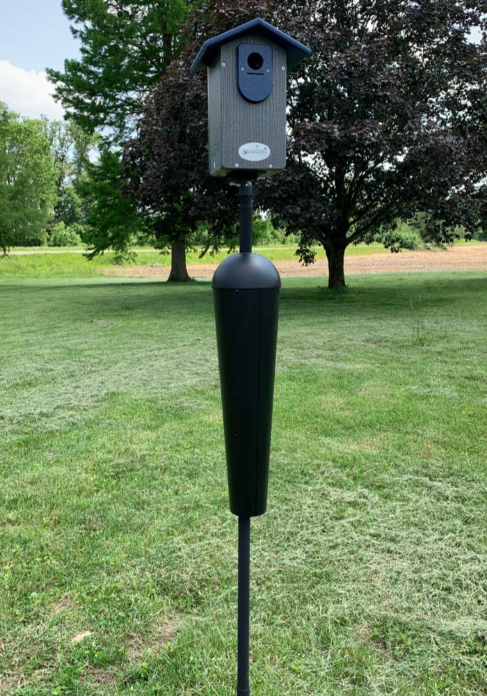 Photograph of a Far-West Wildlife Designs birdhouse on a tall post with a black predator guard in a grassy yard near trees and a distant field showcasing its gray wood-like texture dark blue roof and sleek design