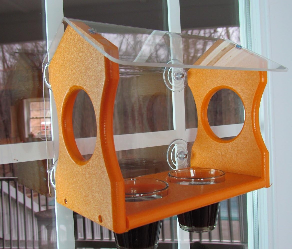 Acrylic bird feeder by unknown artist showing an orange double feeder with clear cups attached to a window with a blurred background showing trees and a house