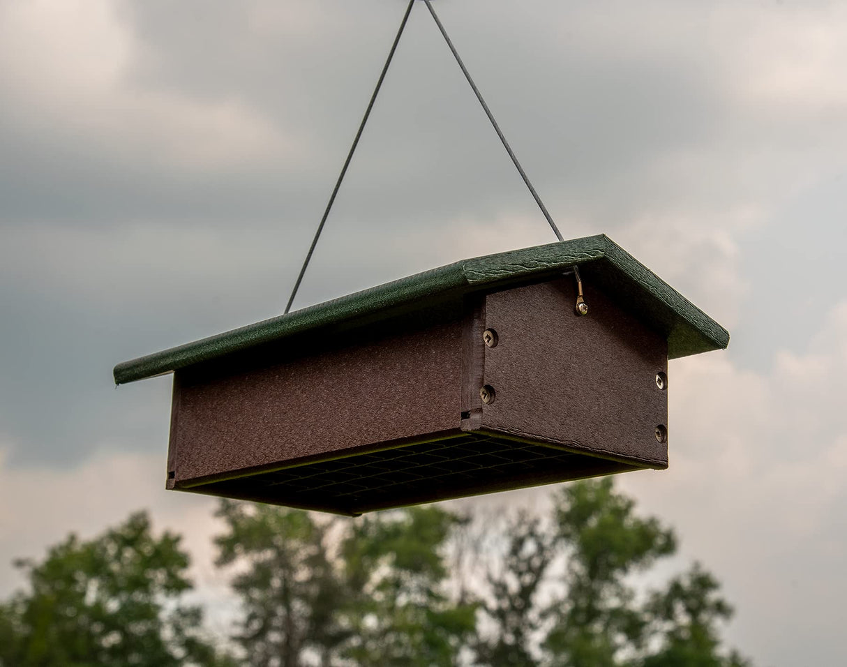 Recycled plastic bird feeder hanging outdoors against a cloudy sky showcasing its dark brown body green roof and visible screws among blurred trees