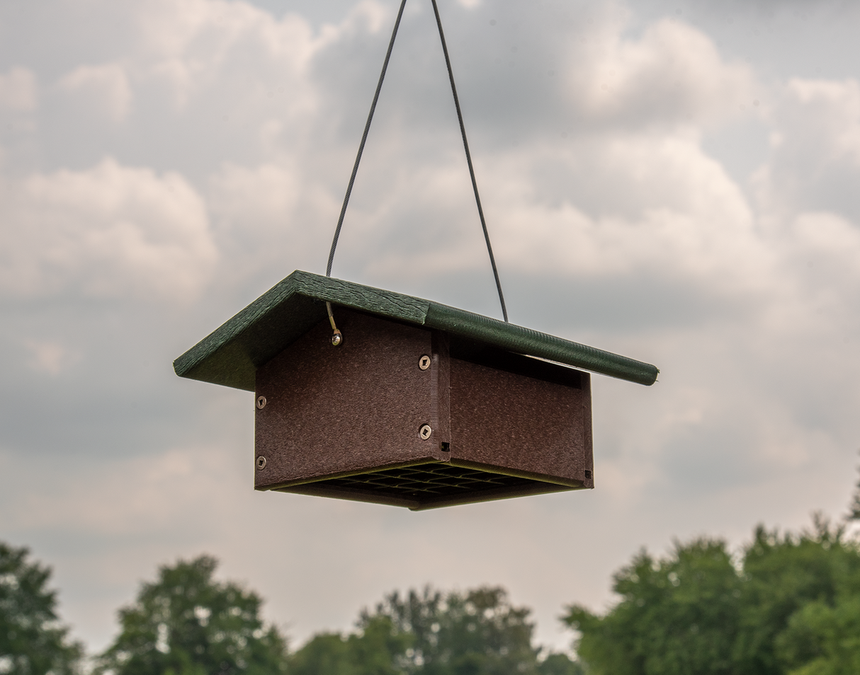 Photograph of a brown and green recycled plastic bird feeder hanging outdoors against a cloudy sky with lush green trees in the background showcasing its dark brown body, a protective green roof, and visible screws.