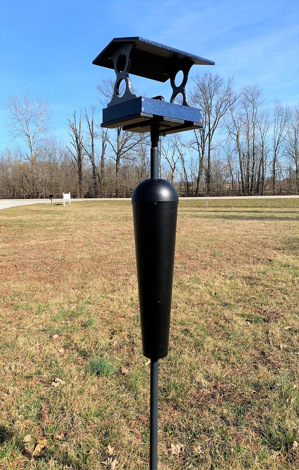 Photograph of a dark blue and black bird feeder with a unique curved roof and a dark post situated in a grassy field under a bright blue sky near bare trees