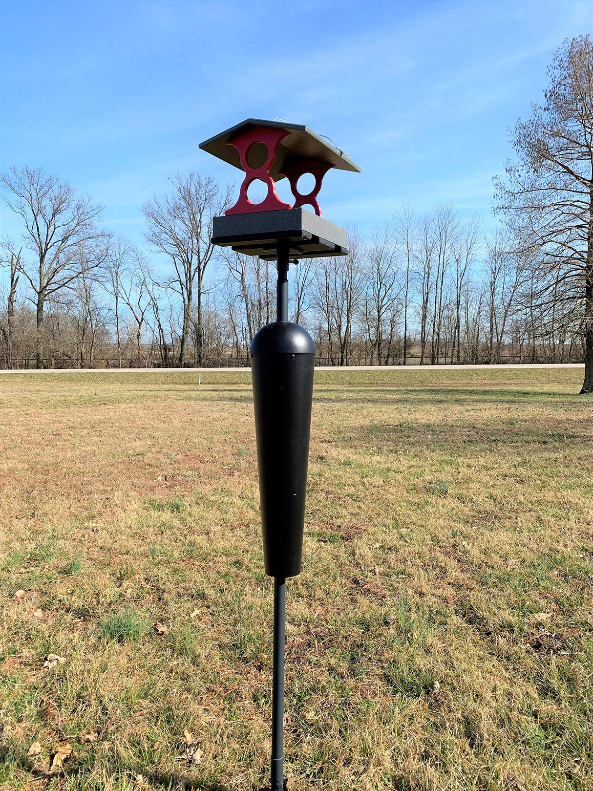 Photograph of a unique bird feeder with a red and gray roof, a black cylindrical hopper, and a tall metal pole situated in a grassy field against a backdrop of bare winter trees and a clear blue sky