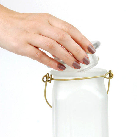 Photograph showing a hand opening a frosted glass jar with a brass wire handle and a clear glass lid featuring mauve nail polish against a white background