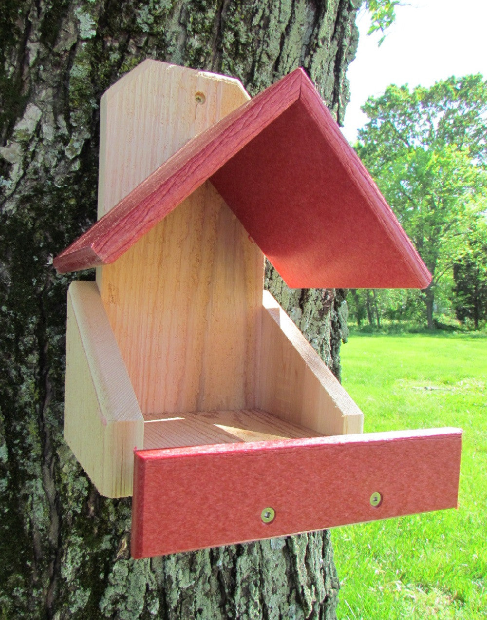 Wooden bird feeder mounted on a tree in a grassy yard featuring a red roof and tray natural wood body and visible screws