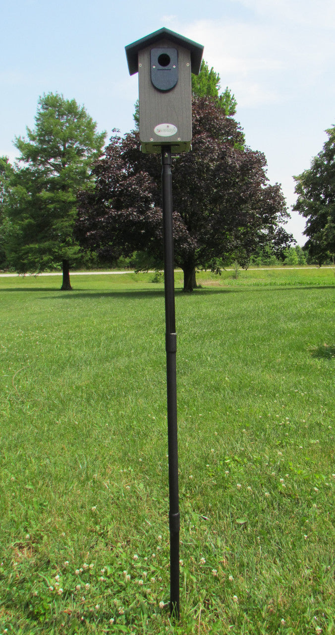 A gray and green birdhouse on a black pole in a grassy yard near trees and a road featuring a dark green roof, a small round entrance hole, and a white logo