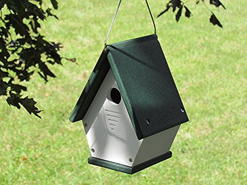 Plastic birdhouse in a garden setting featuring a dark green roof white body and a small entry hole hanging from a tree branch