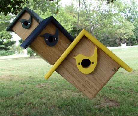 Three handcrafted wooden birdhouses in green yellow and blue hanging outdoors against a backdrop of trees and a grassy lawn featuring whimsical bird silhouettes and visible screws