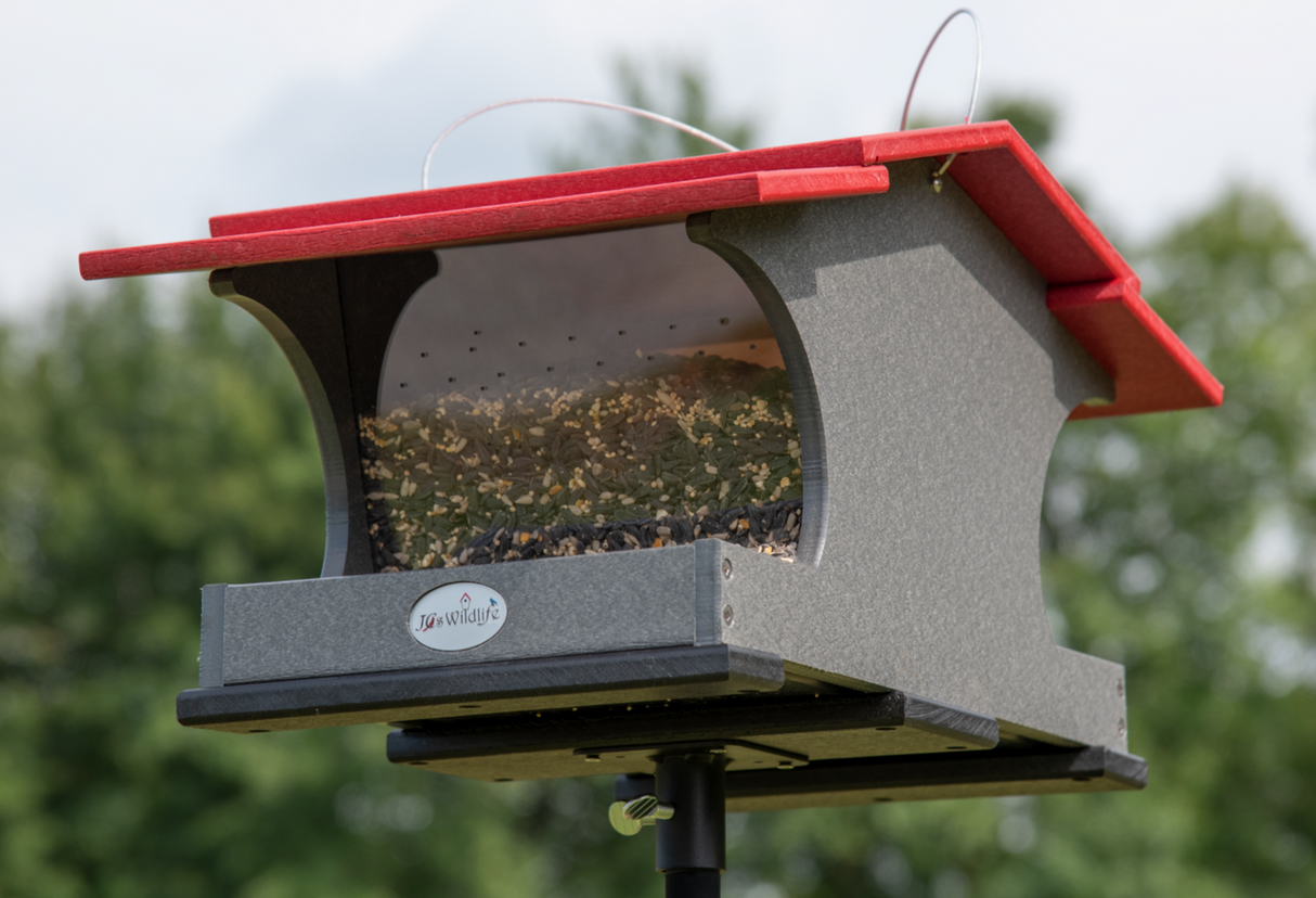 JQ's Wildlife gray and red recycled plastic bird feeder filled with mixed seeds stands on a black metal post against a blurred green background.
