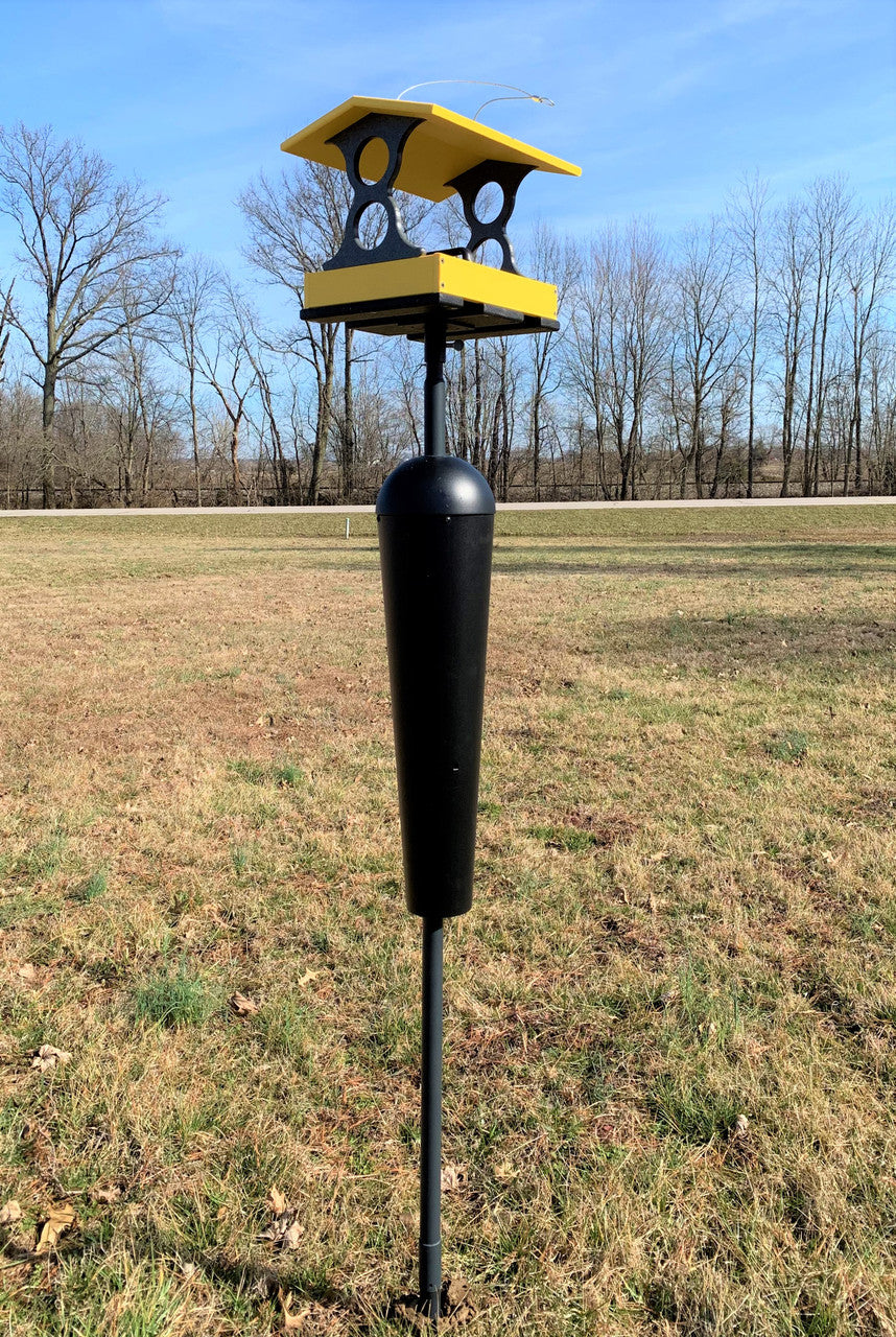 Contemporary bird feeder with yellow and black accents situated in a grassy field near bare winter trees and a distant road