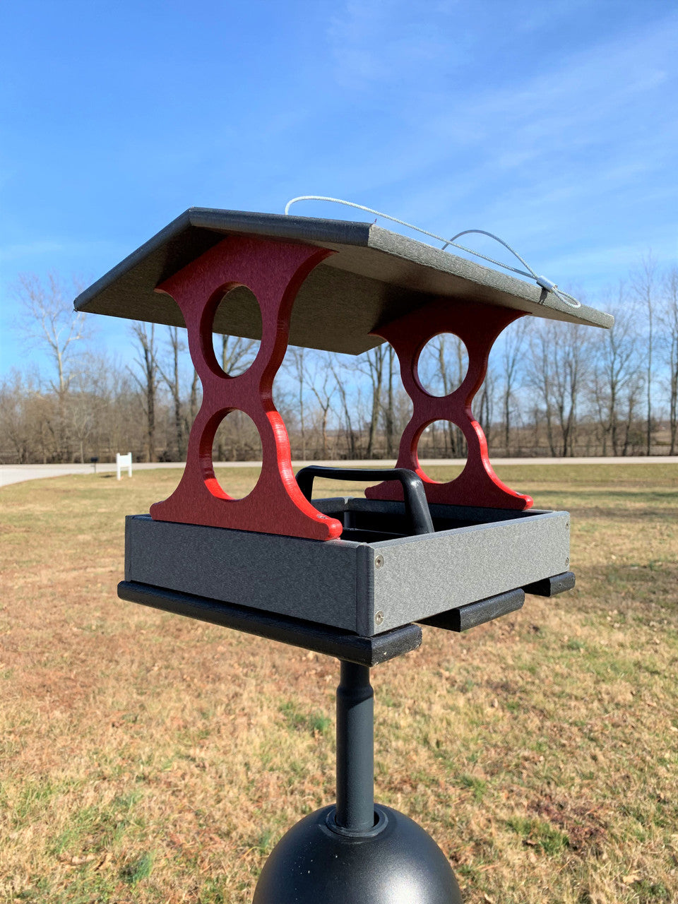 Recycled plastic bird feeder in gray and red featuring a pitched roof and circular cutouts situated outdoors against a backdrop of trees and a clear blue sky
