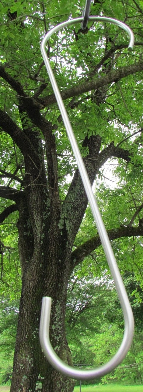 Photograph of a large, shiny metal S-hook hanging from a tree with lichen-covered bark in a verdant landscape featuring a partly visible fence in the background