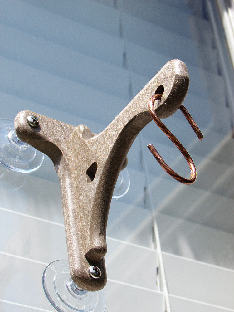 Close-up photograph of a tan plastic suction cup hummingbird feeder with a shiny copper wire hook attached against a light blue window blind background showing visible screws and clear suction cups