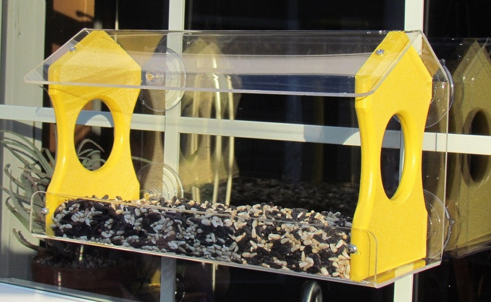 Acrylic and wood bird feeder attached to a window showing mixed birdseed, bright yellow accents, and clear suction cups visible against a windowpane reflecting a blurry outdoor scene