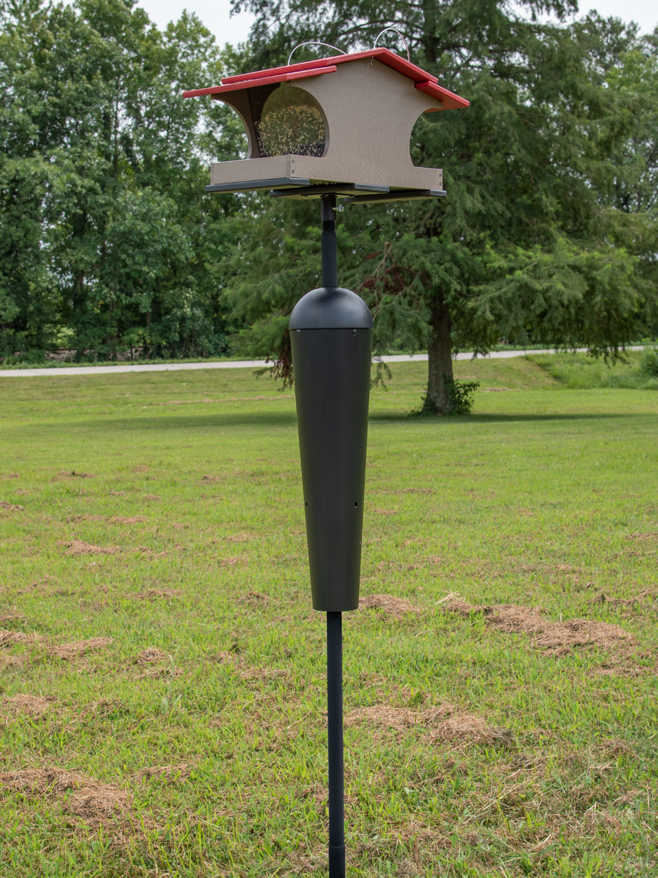 Photograph of a two-tiered bird feeder with a red roof and a black cylindrical squirrel baffle standing in a grassy field near trees and a road showing seed and detailed craftsmanship