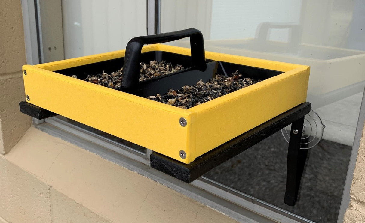 Handmade window bird feeder with bright yellow tray black accents and birdseed on a windowsill.