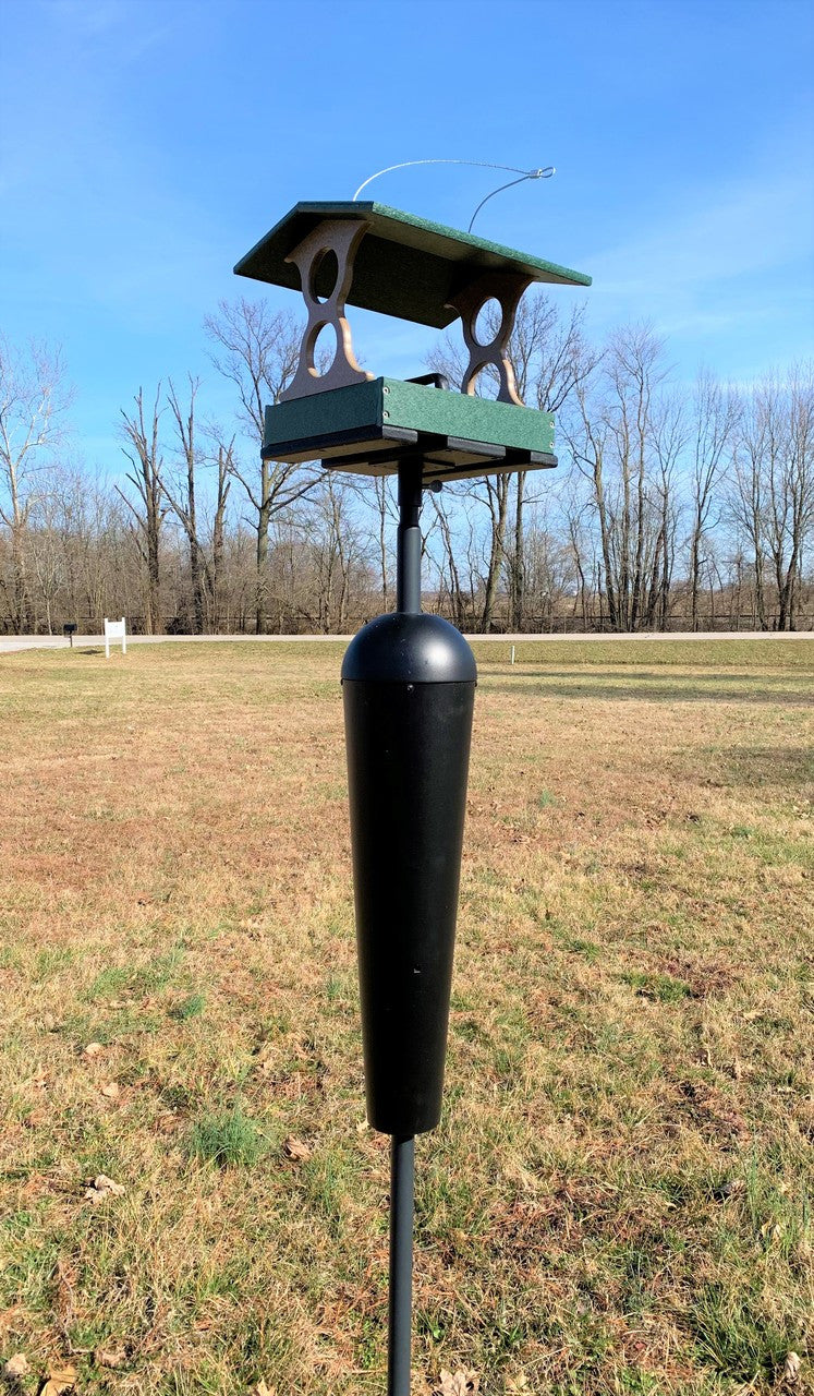 Photograph of a green and brown bird feeder with a black squirrel baffle on a metal pole in a grassy field with bare trees in the background under a bright blue sky