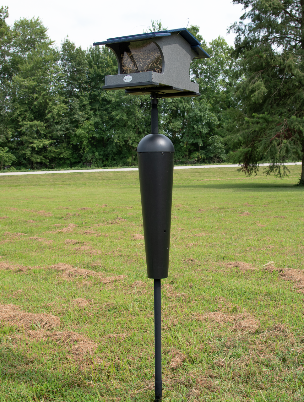 Photograph of a gray and blue bird feeder with a black protective squirrel baffle on a tall black pole in a grassy field near a line of trees