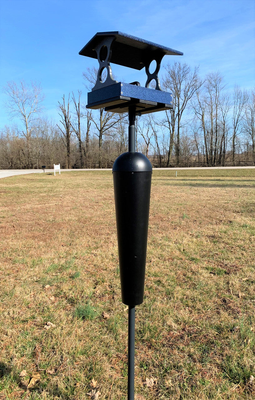 Photograph of a dark blue and black bird feeder with a squirrel baffle set in a grassy field near bare winter trees and a white sign.