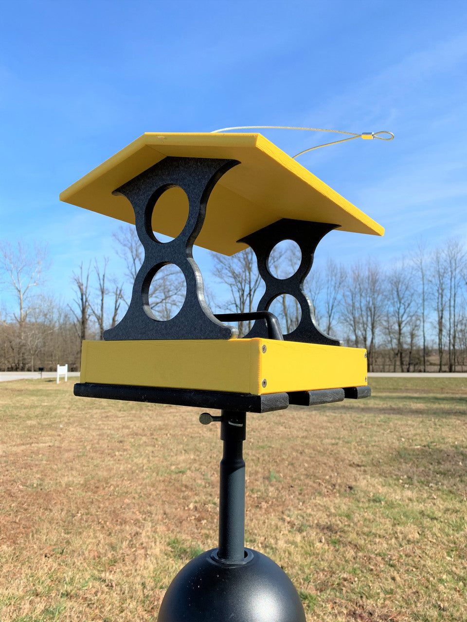 Photograph of a yellow and black bird feeder on a stand in a rural field with trees and a clear blue sky showing a bright yellow tray and decorative cutouts