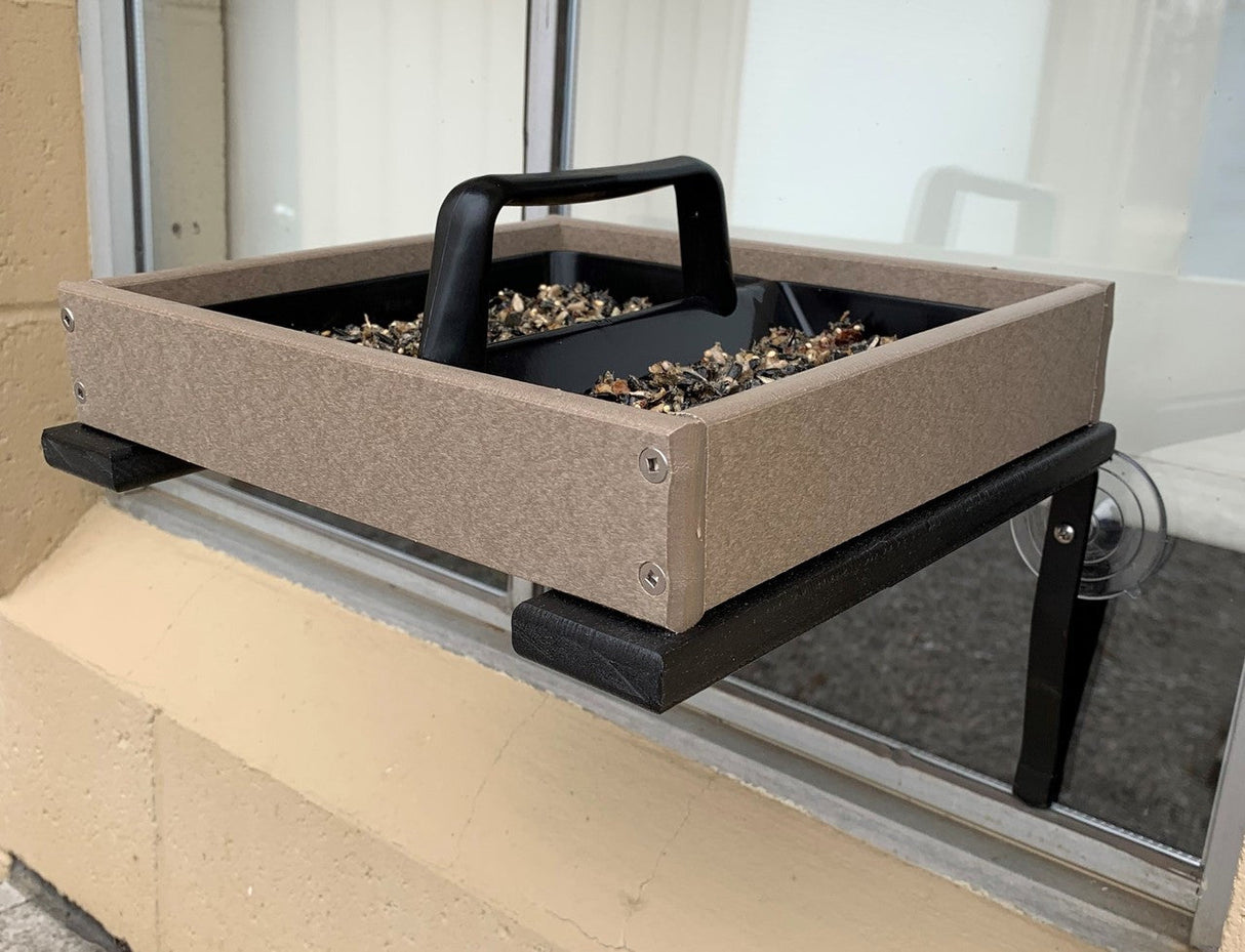 Photograph of a taupe and black recycled plastic window bird feeder with a black handle filled with birdseed attached to a window with suction cups showing visible screws and a textured surface