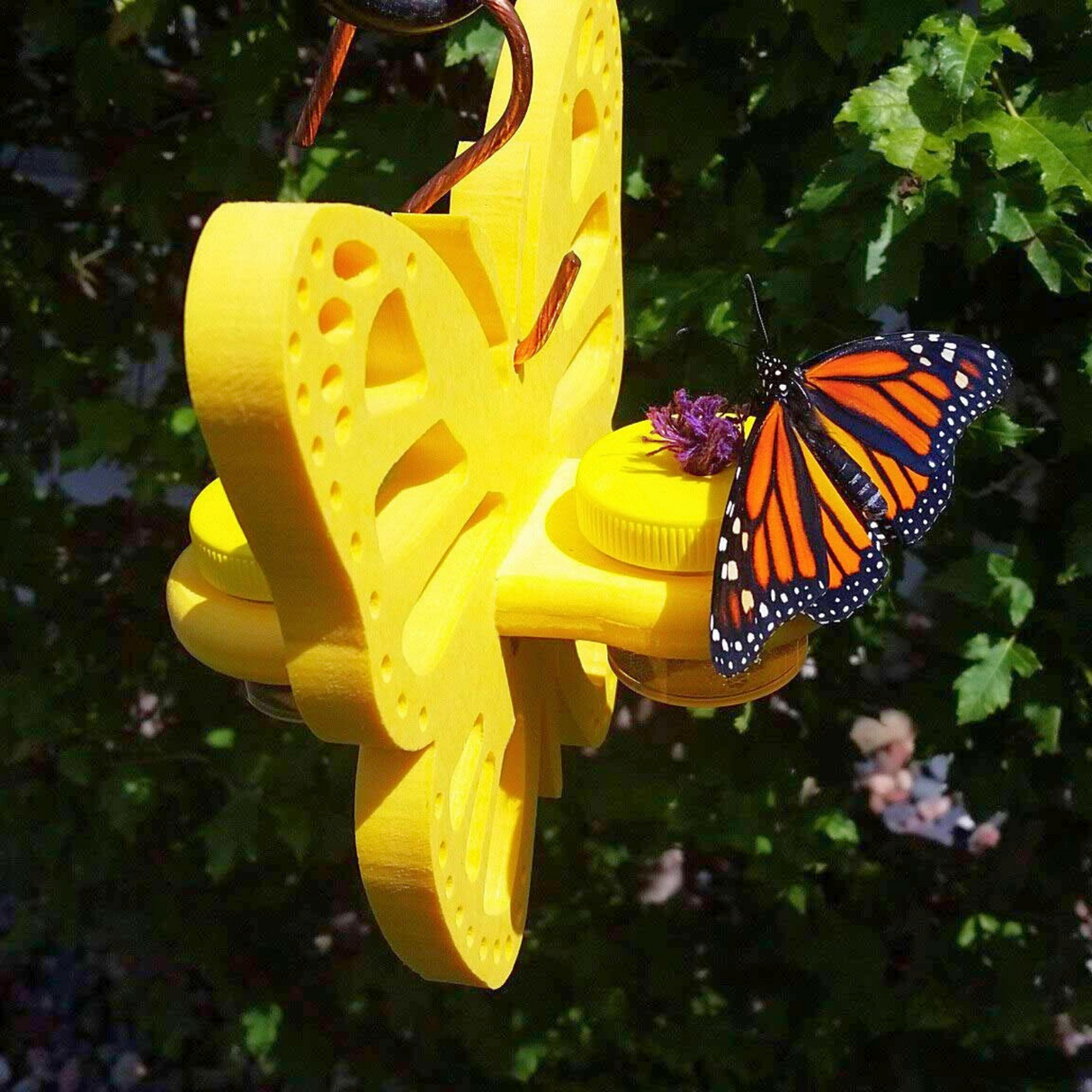 3D printed art, a monarch butterfly feeding on a purple flower atop a bright yellow butterfly-shaped feeder with two small yellow jars against a lush green background