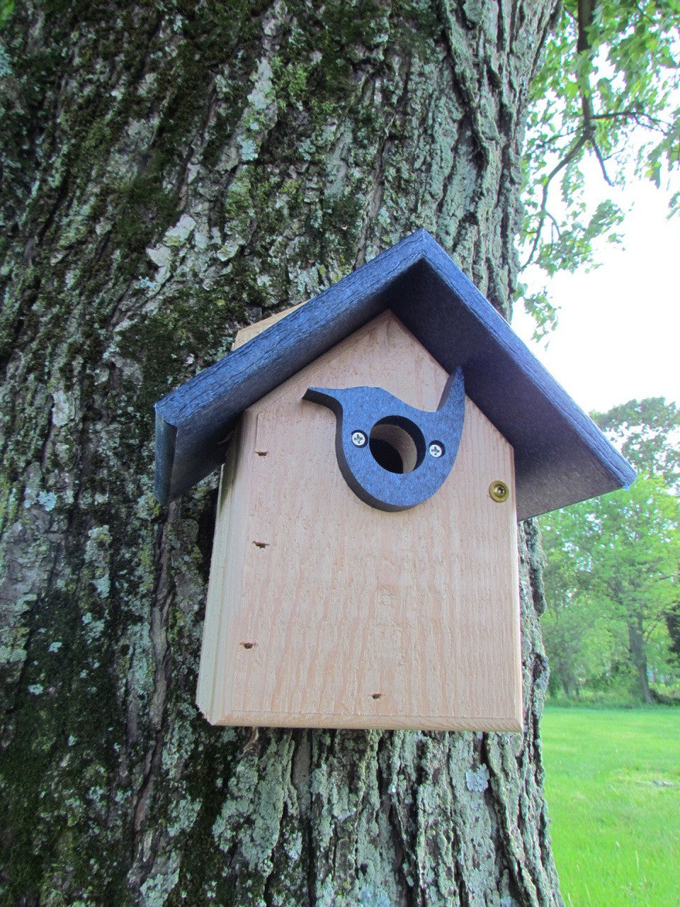 Handcrafted wooden birdhouse with a dark blue roof and bird-shaped entrance on a moss-covered tree in a green field