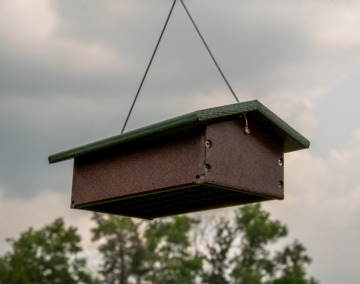 Recycled plastic bird feeder hanging outdoors against a cloudy sky showing dark brown body, green roof, and visible screws