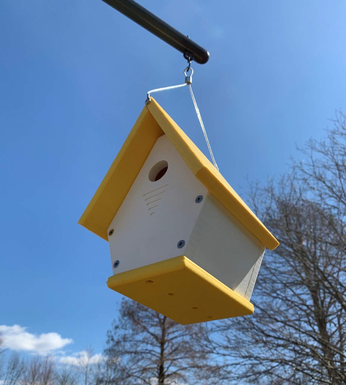 3D-printed birdhouse hanging outdoors against a bright blue sky featuring a cheerful yellow roof and base showcasing  bare trees in the background