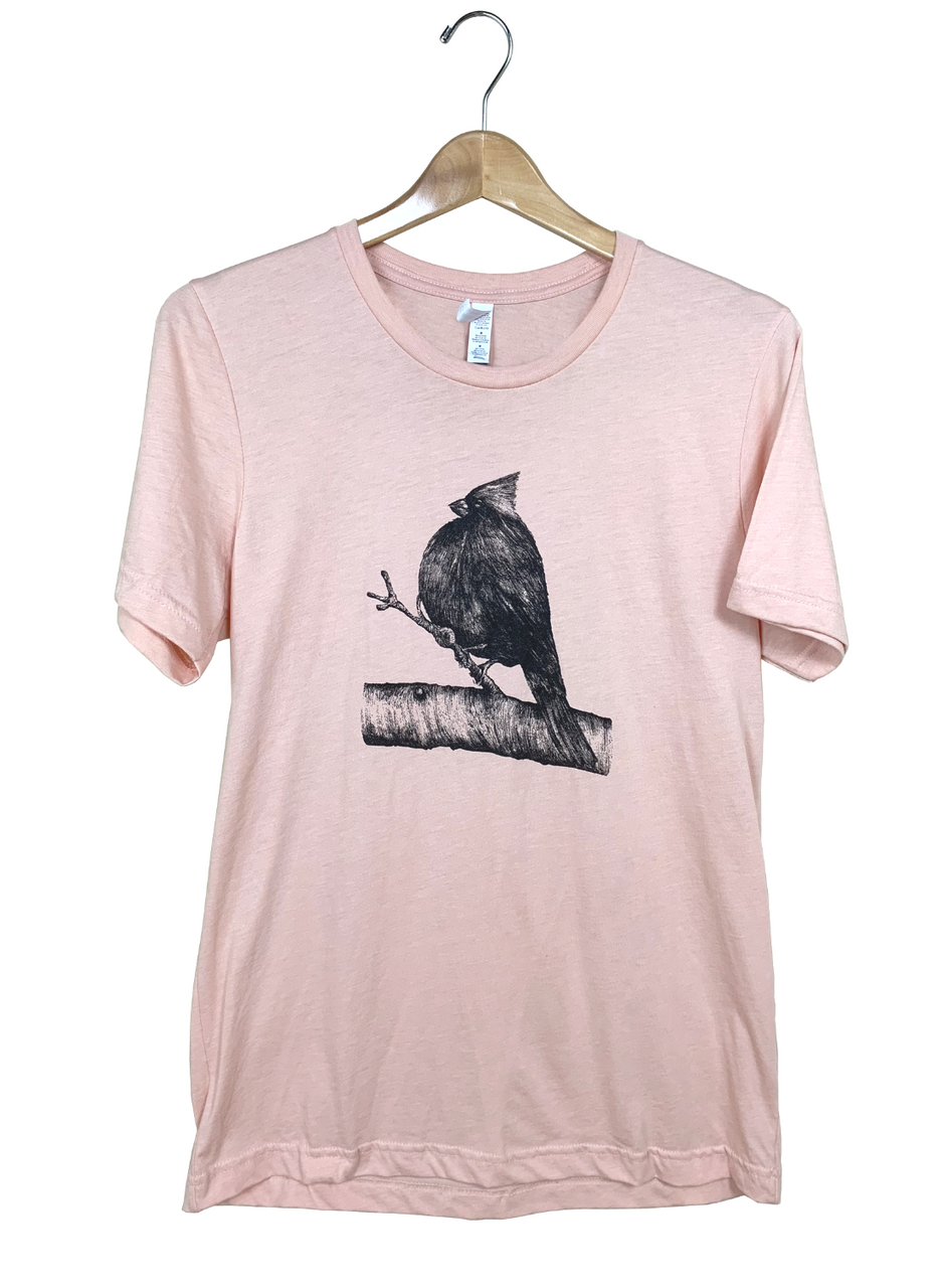 Apparel graphic design featuring a Northern Cardinal perched on a detailed branch with textured bark and delicate twigs against a pale pink t-shirt