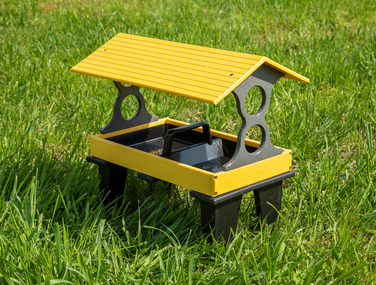 Plastic bird feeder on grass featuring a yellow roof, black legs, and a perforated tray for seed dispensing