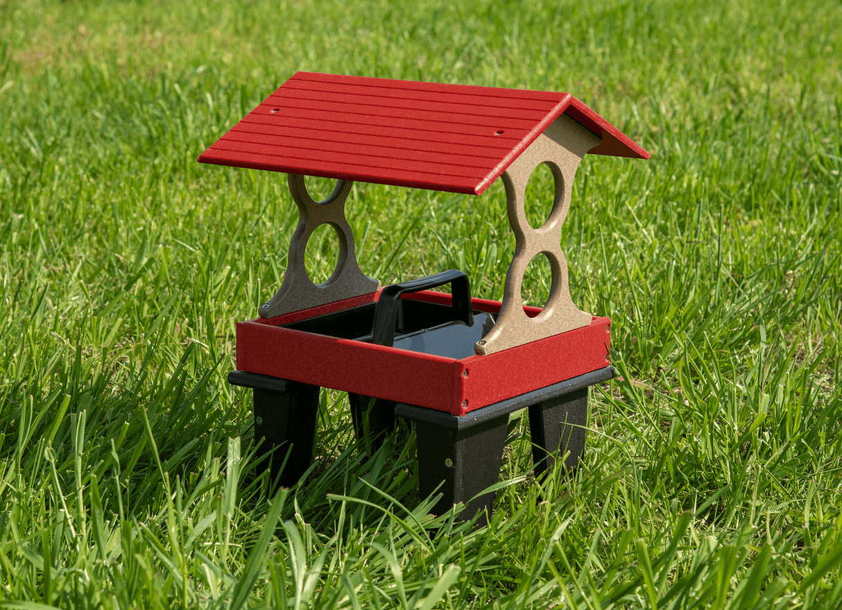 Plastic recycled bird feeder on green grass with a red roof and taupe supports featuring a black tray and carrying handle